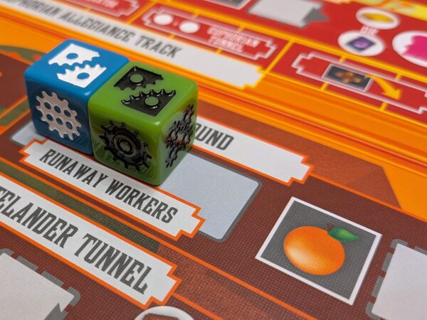 Due dadi con icone di ingranaggi, uno blu e uno verde, posizionati sul tabellone di un gioco da tavolo